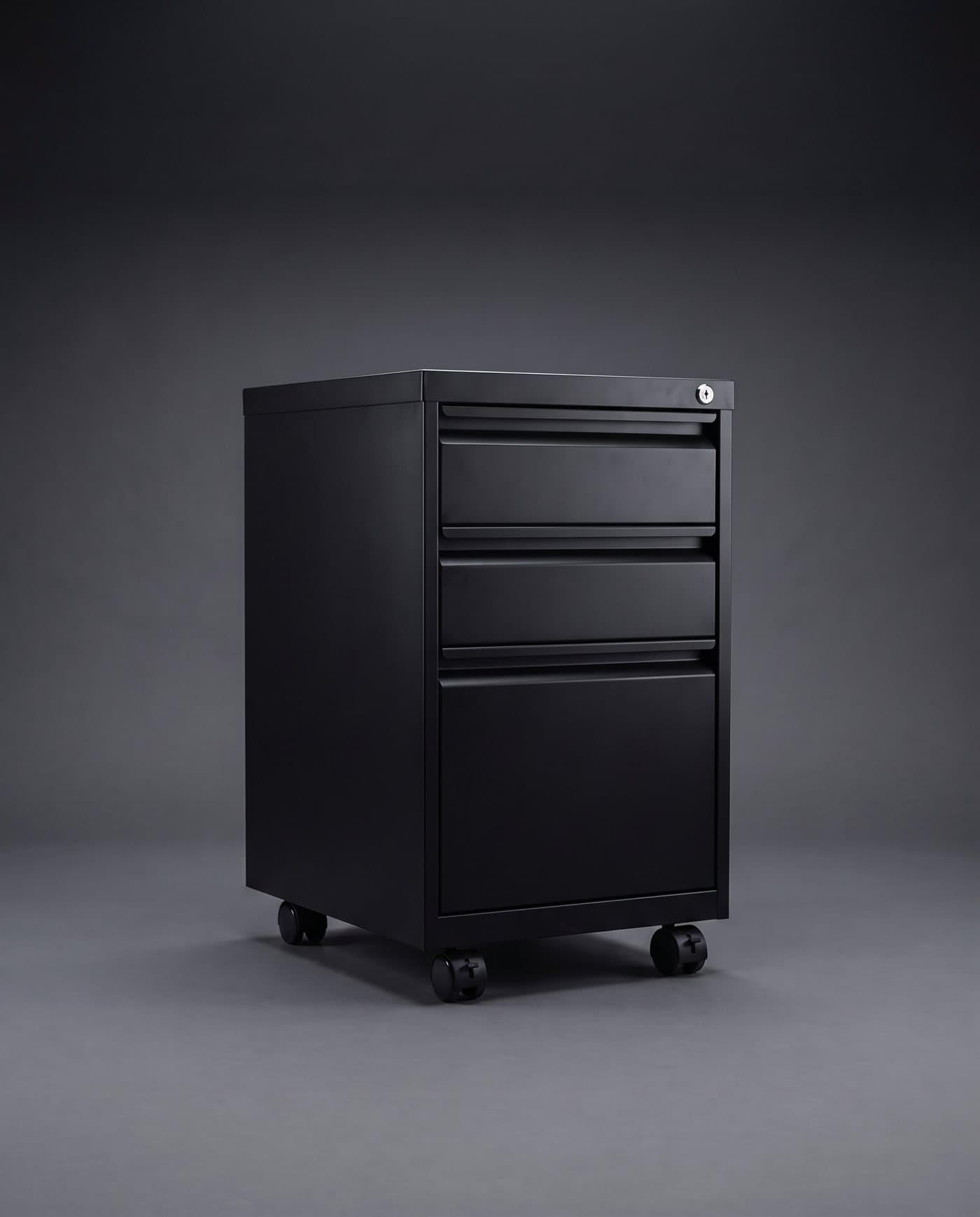 Caisson à trois tiroirs en acier noir mat sur roulettes, serrure en haut à droite, sur fond anthracite