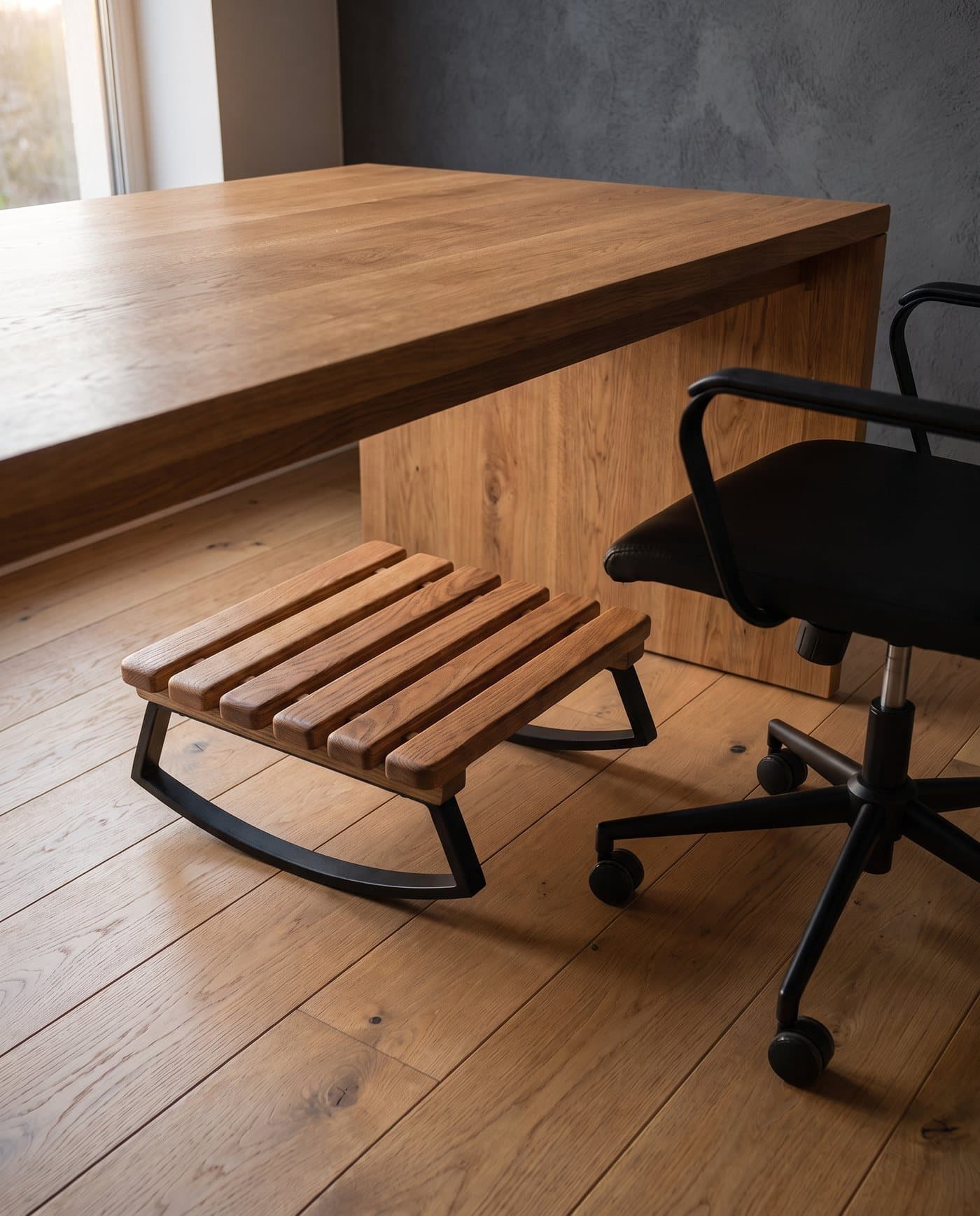 Le repose-pieds Idle posé sur un parquet sous un bureau en chêne, à côté du piètement d'une chaise